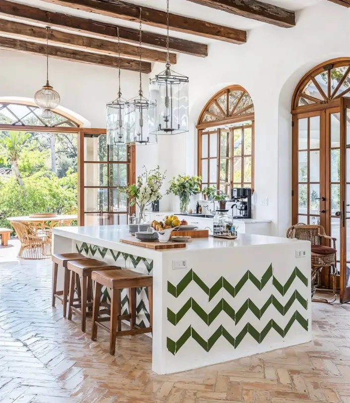 A Mediterranean kitchen interior with rustic ceiling beams, a chevron-tiled island, and arched wooden doors.