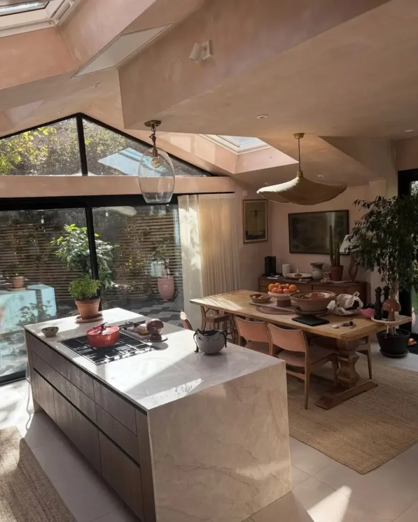 A warm minimalist kitchen with blush walls, a marble island, and a wood dining table bathed in sunlight.