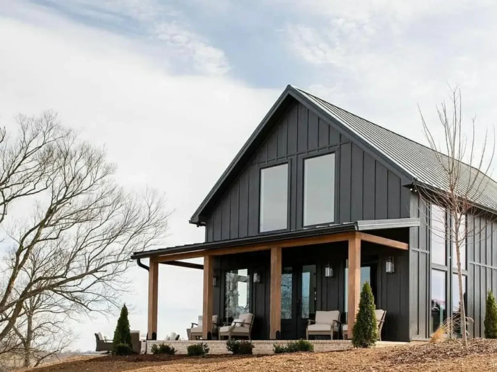A modern black farmhouse on a hill with a large wraparound porch supported by natural wood posts.