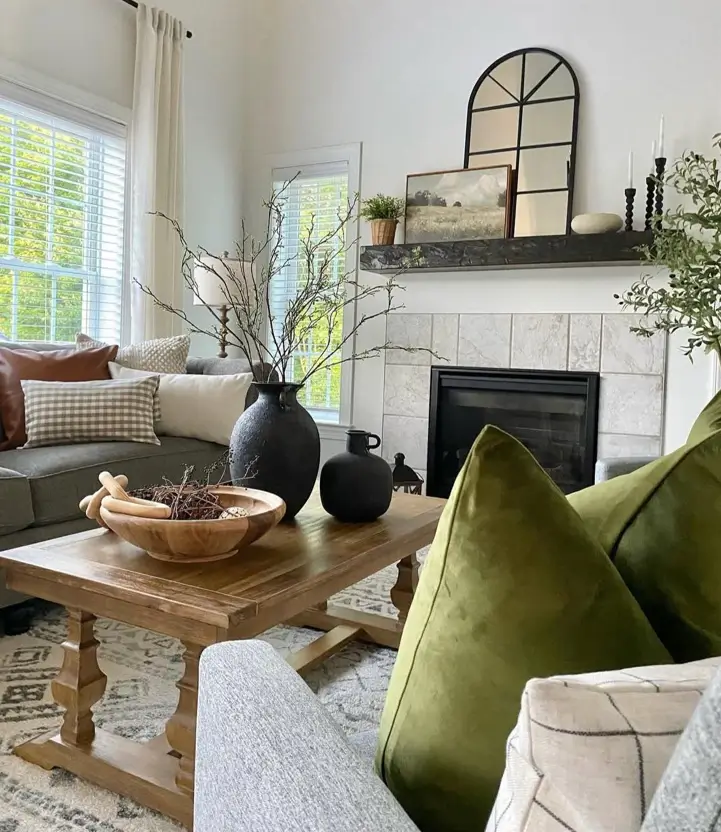 A neutral living room with a gray sofa, olive green pillows, and a rustic wood coffee table in front of a fireplace.