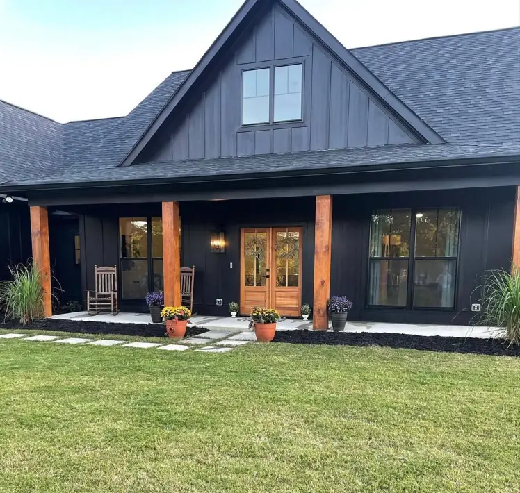 A modern black farmhouse with a deep covered porch, thick wood posts, and a natural wood double door.
