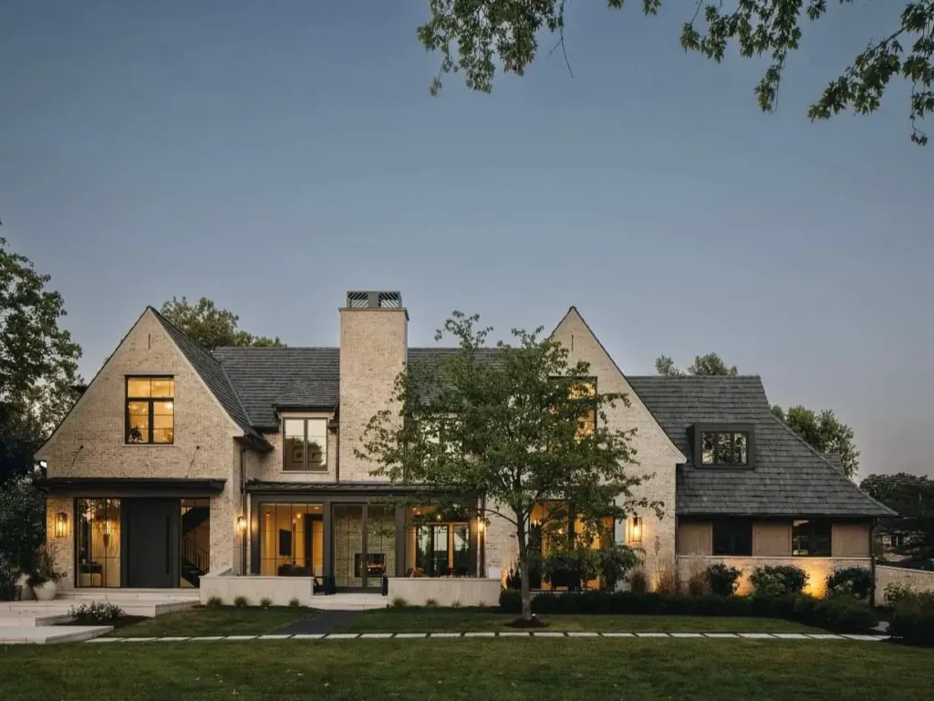 A large beige brick house exterior with a dark slate roof and black-trimmed windows, illuminated at dusk.