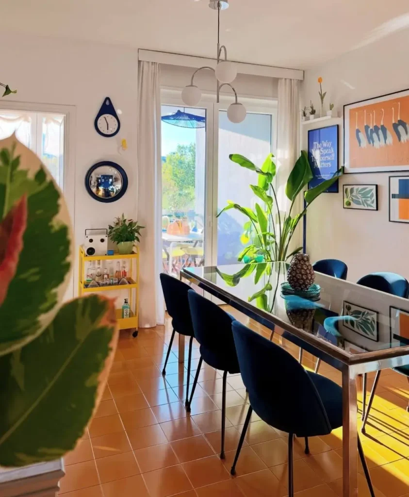 A bright, cozy small apartment dining room with blue velvet chairs, a yellow bar cart, and colorful art creating a vibrant interior.