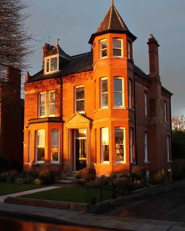 A classic red brick modern Victorian house with a corner turret, glowing in the warm light of sunset.