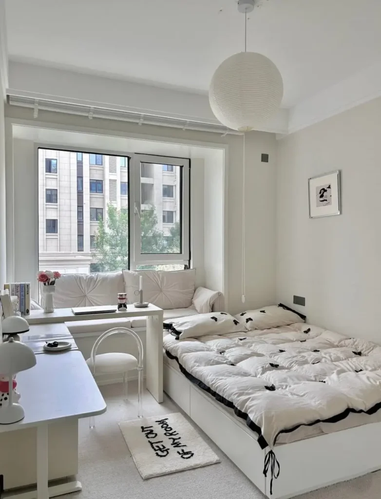 A serene, cozy small apartment bedroom featuring a monochromatic white interior, a built-in window seat, and a tidy workspace.