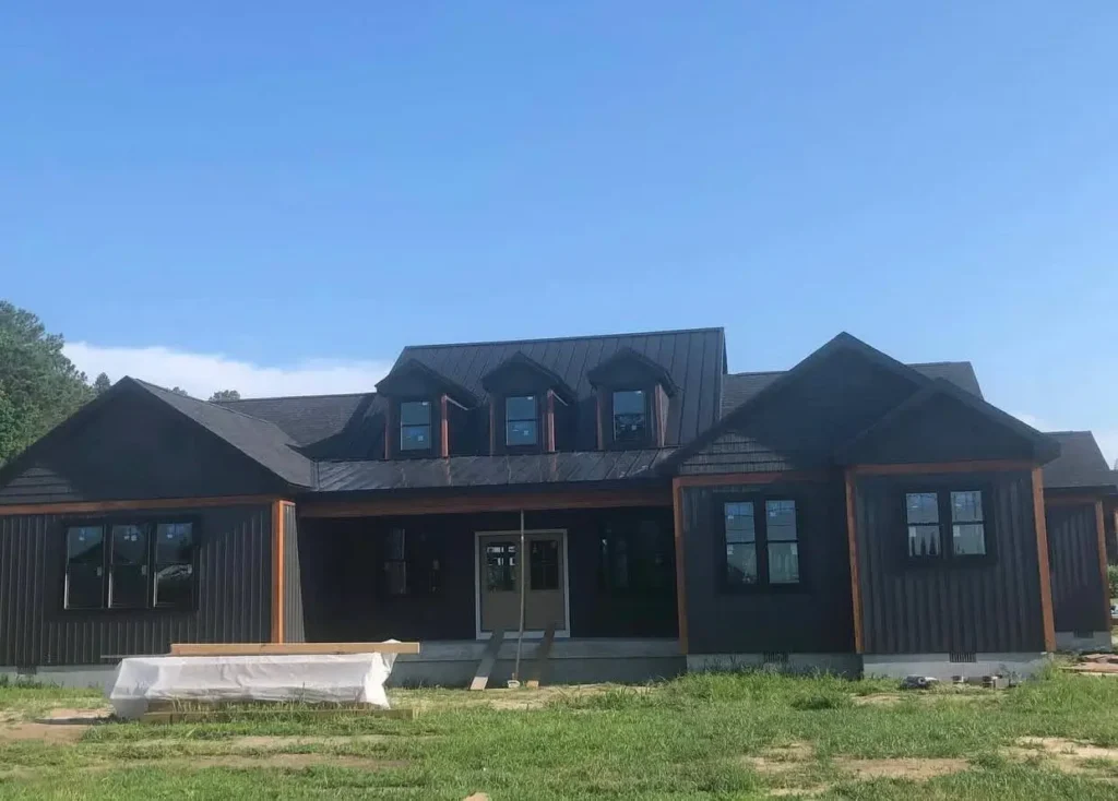 A modern black ranch-style farmhouse under construction with a row of dormers on a metal roof and cedar shake accents.