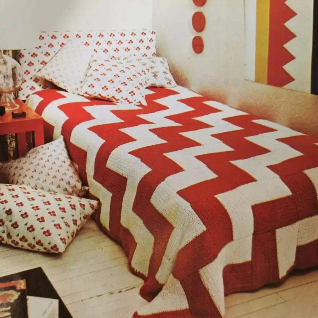 A bed with a bold red and white chevron knit blanket and floral pillows, showing a 70s bedroom idea.