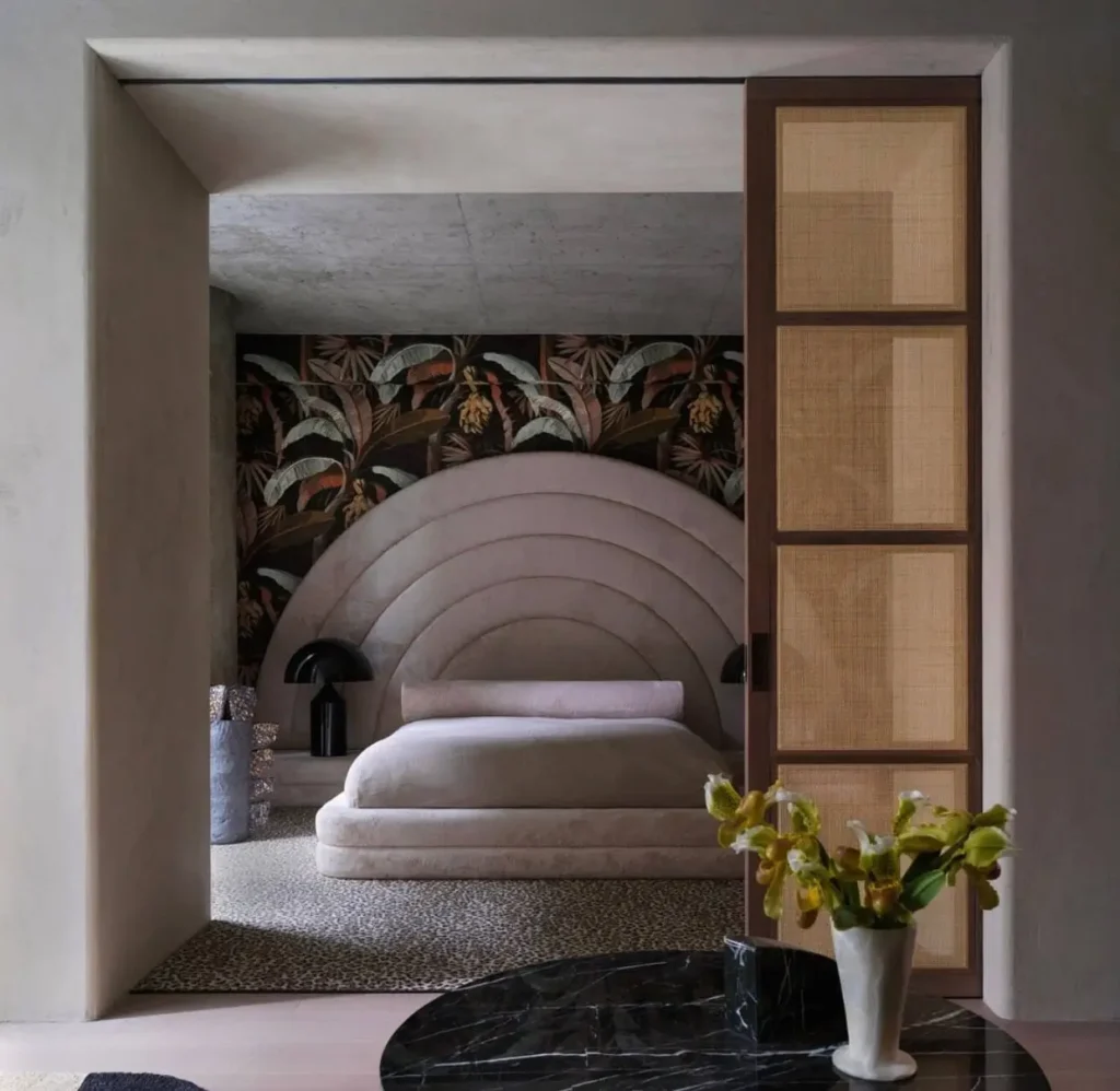 A bedroom with 70s interior design ideas, featuring a large arched headboard, tropical wallpaper, and a cane door.