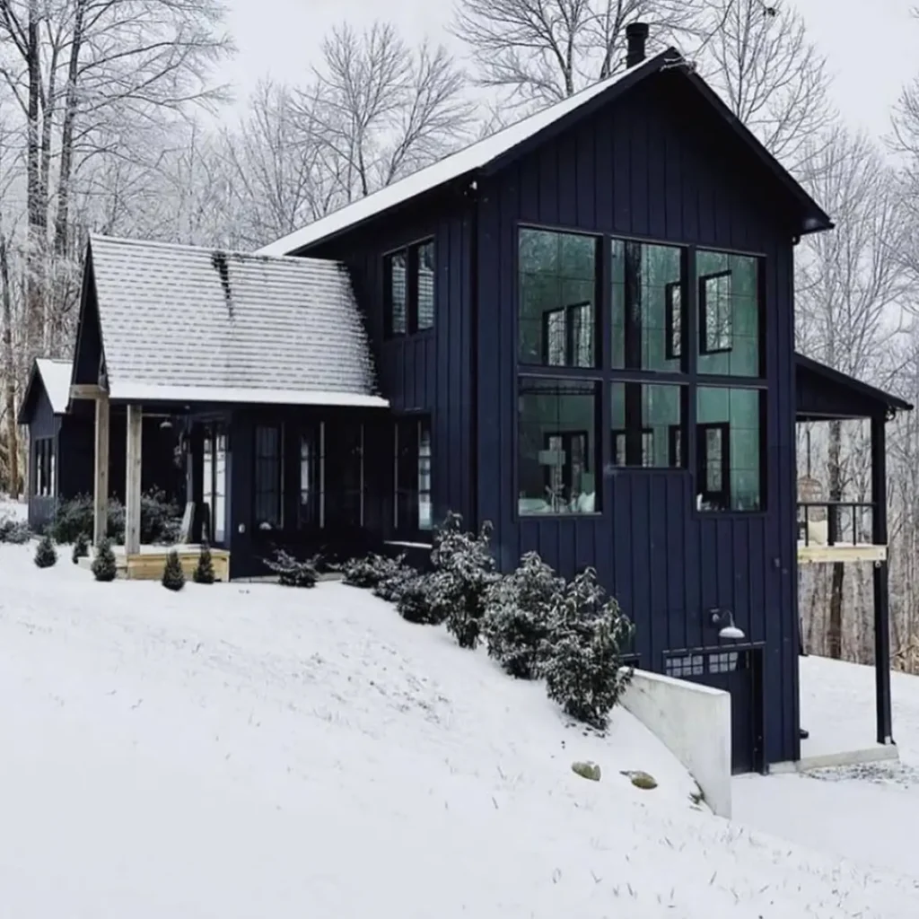 A modern black farmhouse on a sloped, snow-covered hill with a large wall of windows and a walk-out basement.