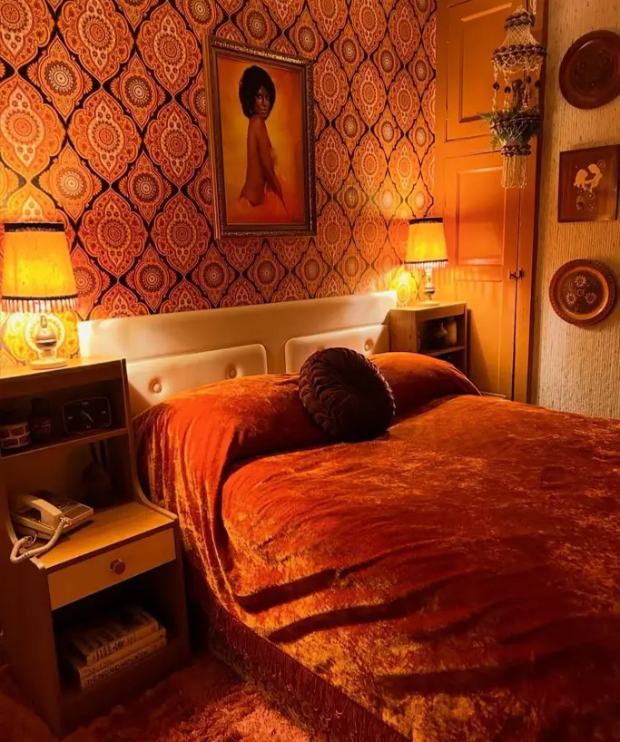 A moody 70s bedroom decor with rust-orange crushed velvet bedding and dark damask wallpaper.