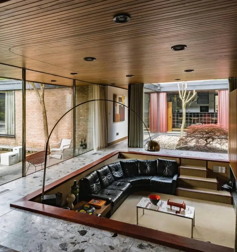 A sunken living room, a key 70s interior design idea, with a black leather sectional, terrazzo floor, and wood ceiling.