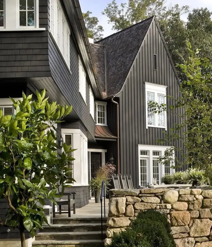 A dark farmhouse exterior with crisp white trim, mixed shingle and vertical siding, and a rustic stone retaining wall.