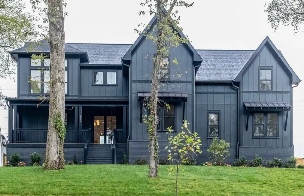 A modern black farmhouse with a monochromatic exterior, including painted black brick, set among mature trees.