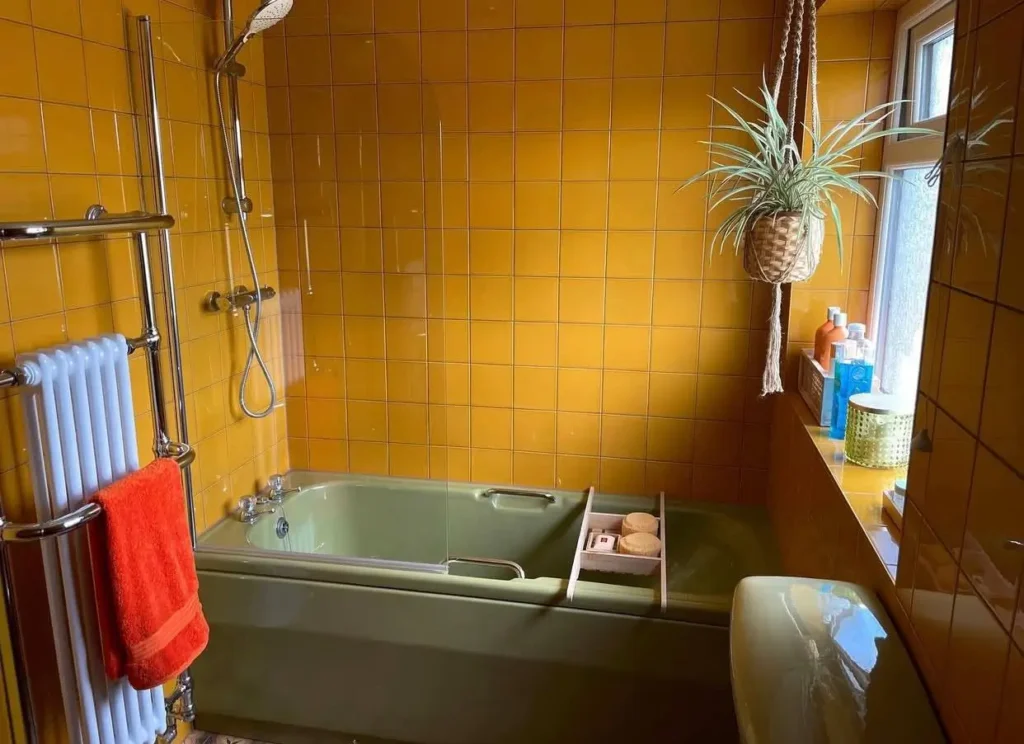 A 70s bathroom with an avocado green tub and toilet set against a backdrop of harvest gold wall tiles.