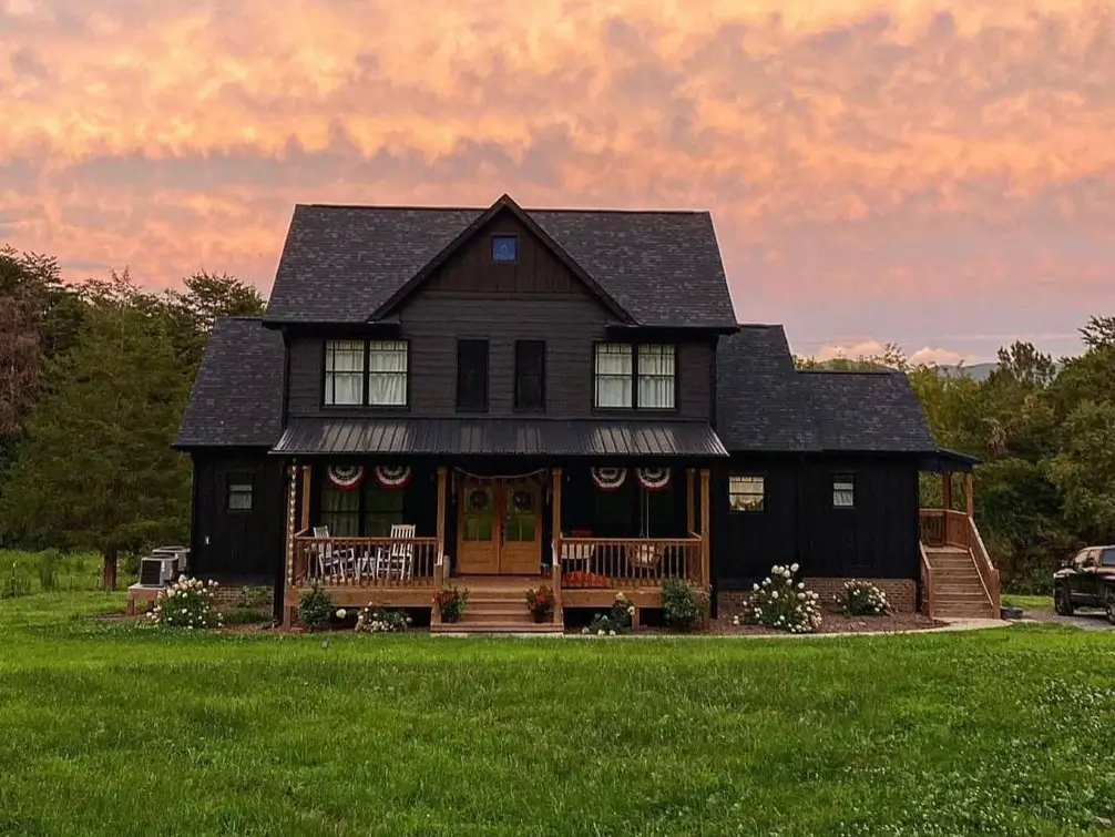 A classic two-story black farmhouse with a full front porch and wood accents glowing at sunset.