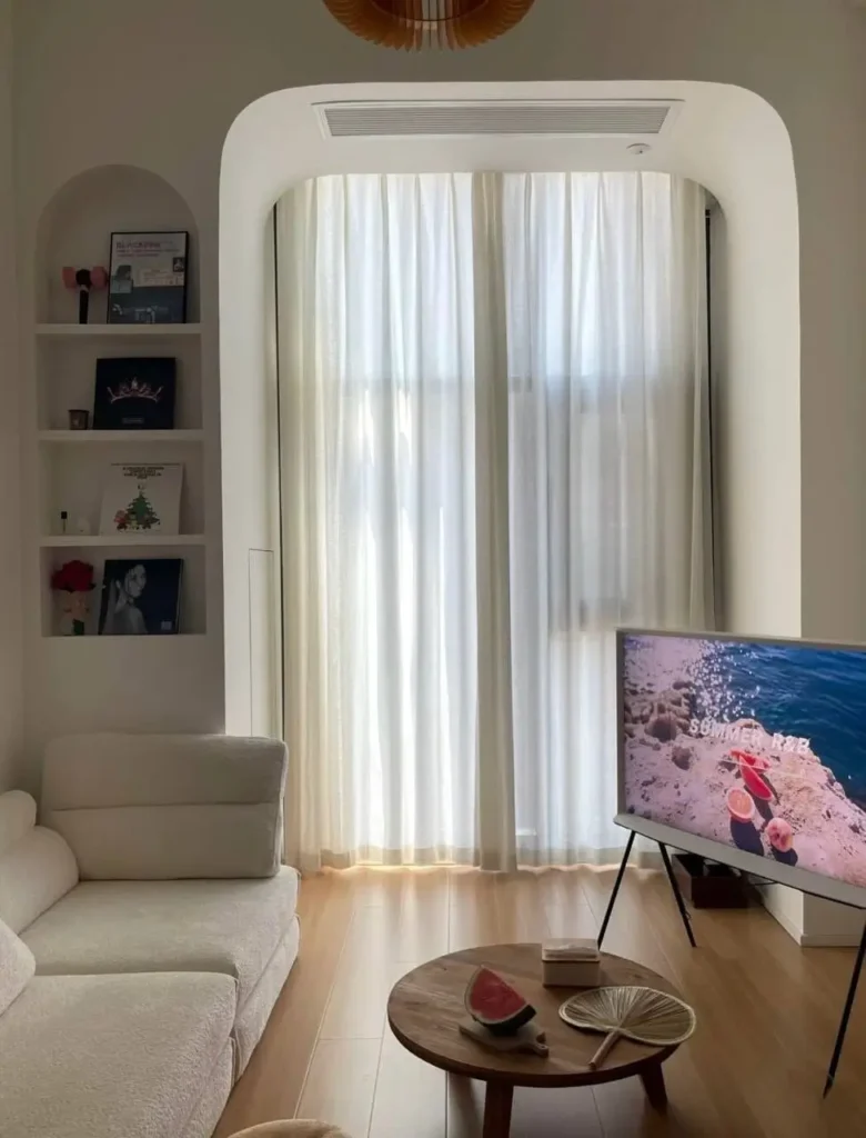 A cozy small apartment living room featuring an arched window with sheer curtains and matching arched shelving for a soft interior design.