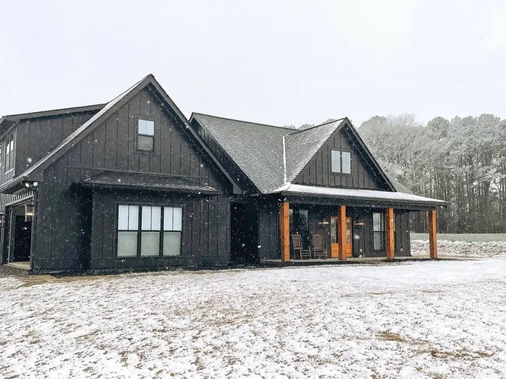 A modern black farmhouse with a wood porch, covered in a light layer of snow during a snowfall.