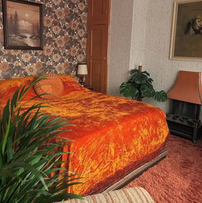 A retro 70s bedroom with orange crushed velvet bedding, dark floral wallpaper, and a shag carpet.