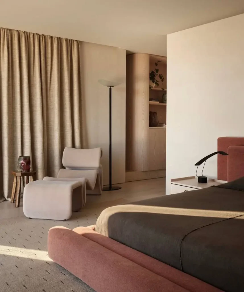 A serene minimalist bedroom with a dusty rose bed, a sculptural beige lounge chair, and long neutral curtains.
