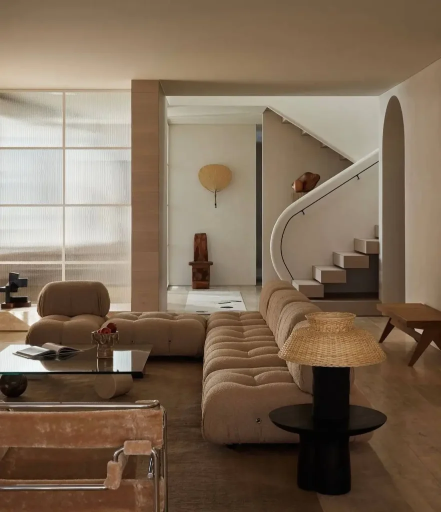 Warm minimalist living room with a curved staircase, tufted beige sofa, and a fluted glass wall.