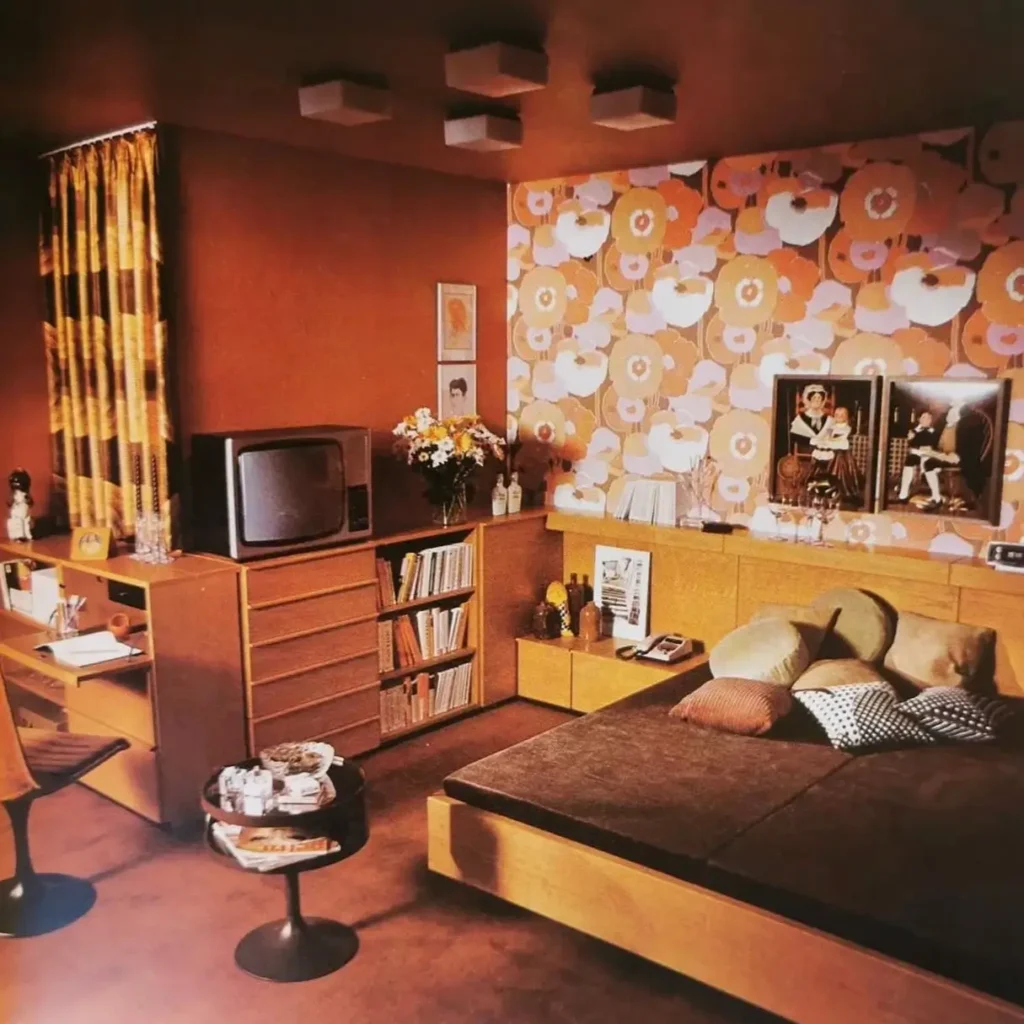 A retro bedroom with bold floral wallpaper, a wood platform bed, and a matching desk unit.