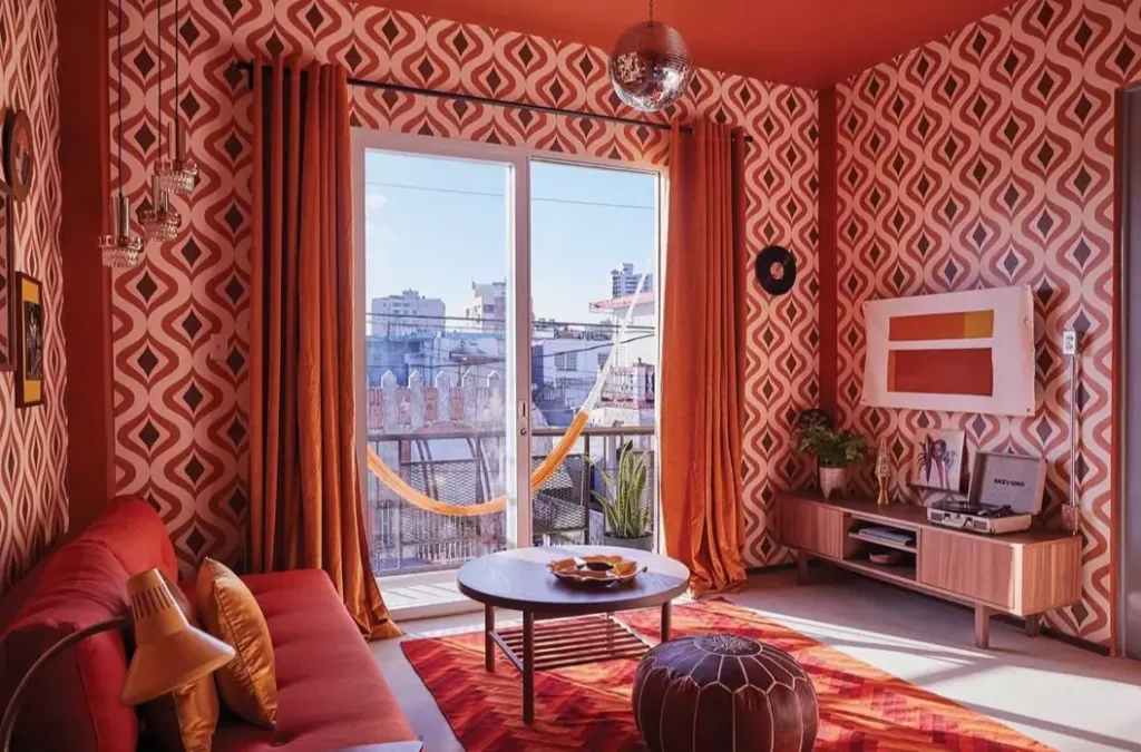 A monochromatic 70s living room with rust-colored patterned wallpaper, matching curtains, and a red sofa.