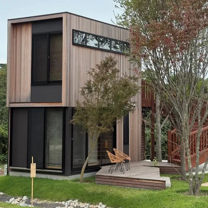 A two-story modern minimalist house with natural wood siding and large black-framed windows, nestled among trees.