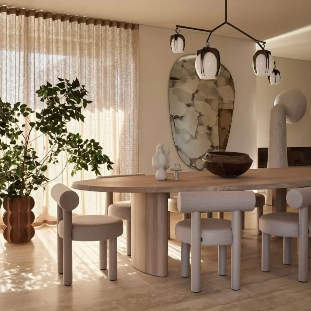 Minimalist dining room with a large wood table, sculptural upholstered chairs, and a modern chandelier in dappled sunlight.
