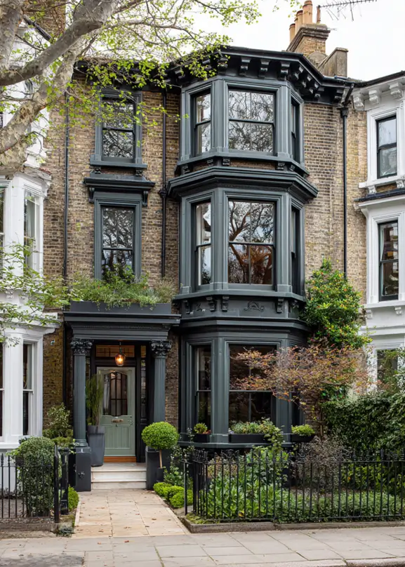 A brick modern Victorian townhouse with dark gray-green bay windows and a light sage green front door.