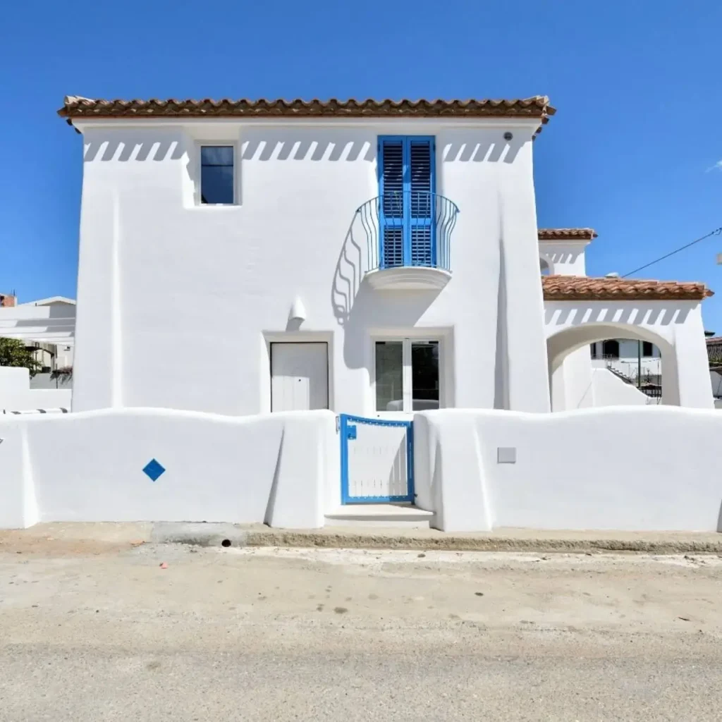 A white Mediterranean house with a terracotta roof and bright blue accents on the shutters, balcony, and gate.