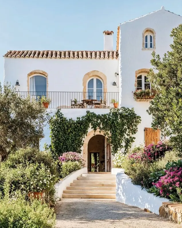 A white Mediterranean villa with climbing vines framing an arched entryway and a garden-lined staircase.