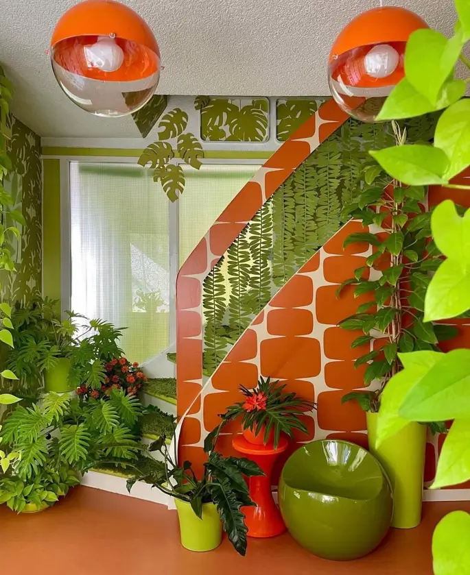 A vibrant room showcasing 70s interior design styles with orange and green graphic wallpaper, plentiful houseplants, and a green pod chair.