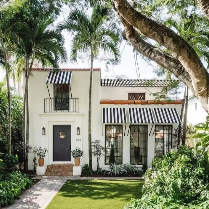 A white Mediterranean house with bold black-and-white striped awnings and lush palm trees.