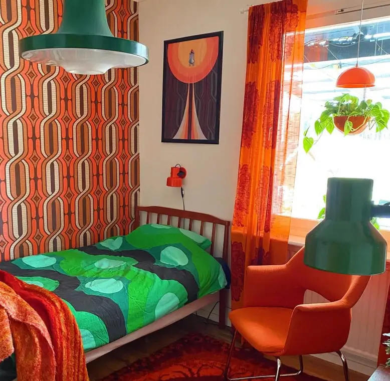 A bedroom with bold 70s geometric wallpaper in orange and brown, paired with a green comforter and an orange chair.