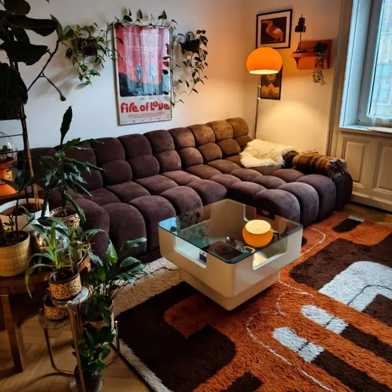 A 70s living room with a large, brown modular sofa, a graphic shag rug, and many houseplants.