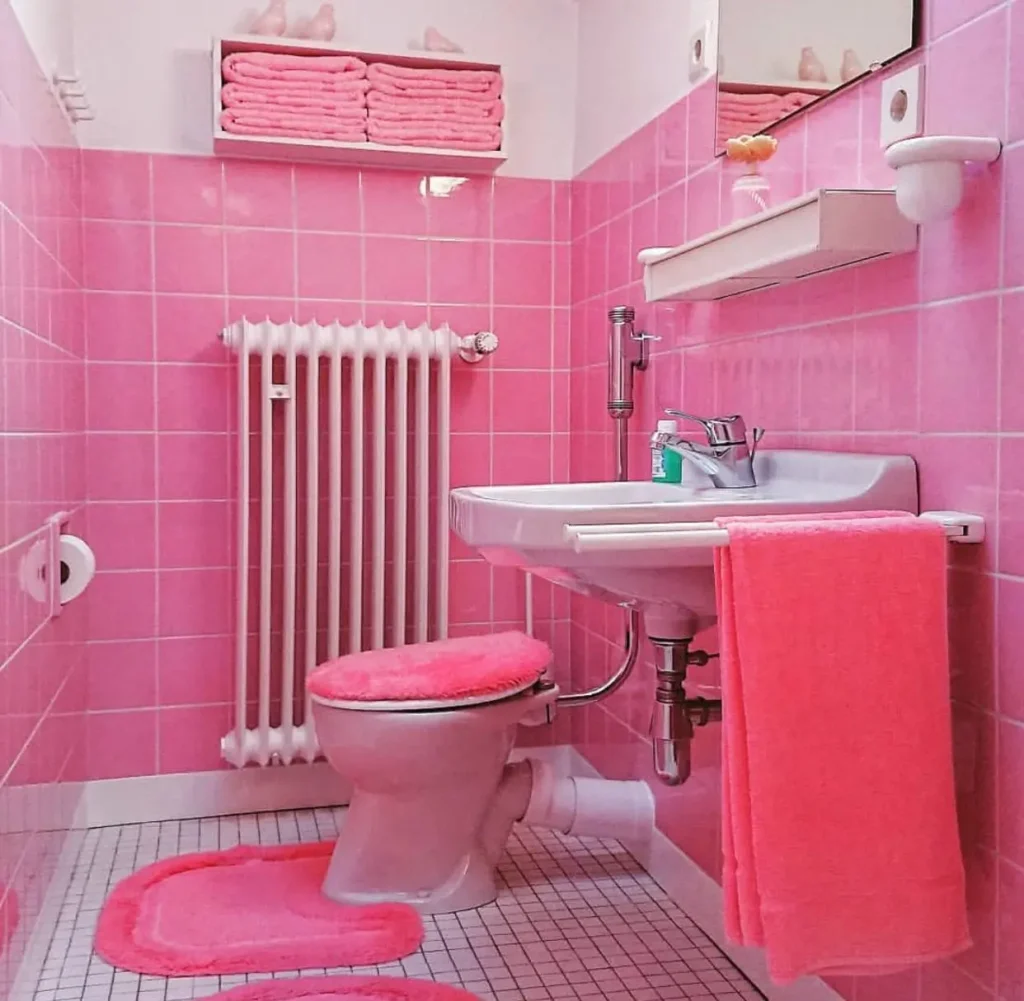 A retro 70s bathroom decorated entirely in saturated pink tile, with matching pink plush toilet covers and rugs.