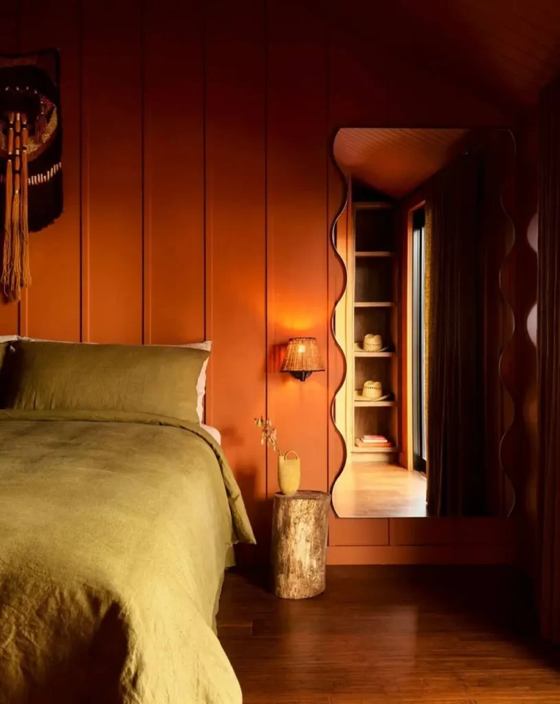 A bedroom showcasing 70s interior design styles, featuring rust-colored wood paneling, olive bedding, and a large wavy mirror.
