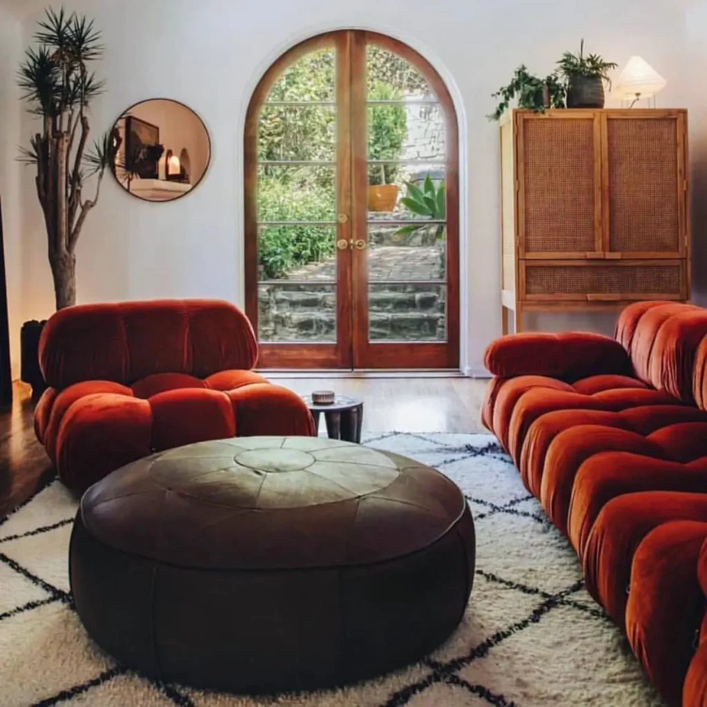 A 70s living room design with a curved, rust-orange modular sofa, a round ottoman, and an arched doorway.