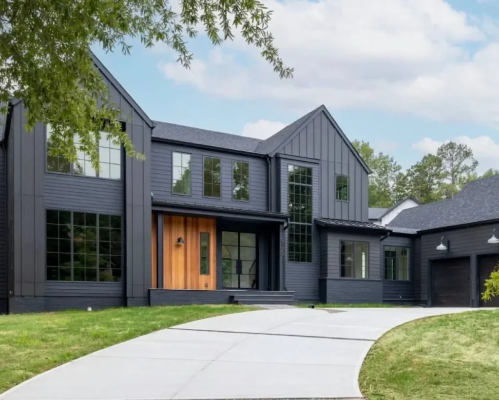 A modern black farmhouse with a recessed natural wood entryway and a long, curved driveway leading to the house.
