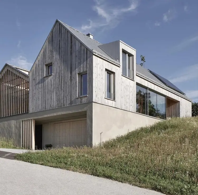 A modern gabled house in Austria by HPSA Architects with weathered wood siding built into a hillside.