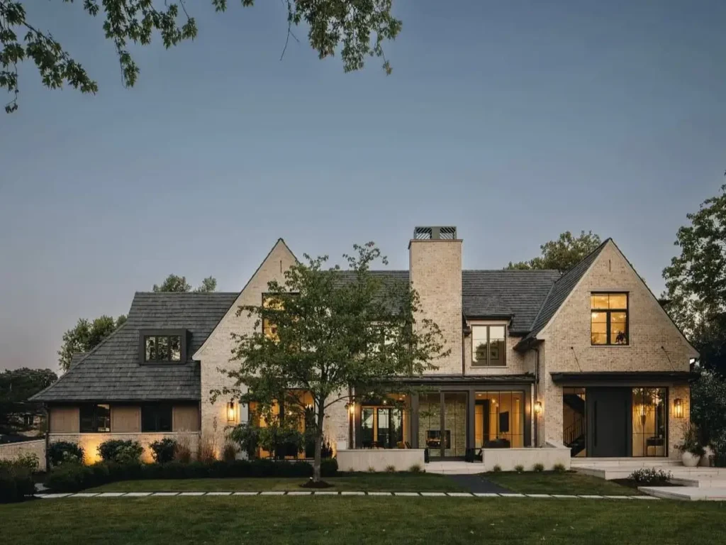 A large beige brick and black house exterior with a dark slate roof and warm interior lighting shown at dusk.
