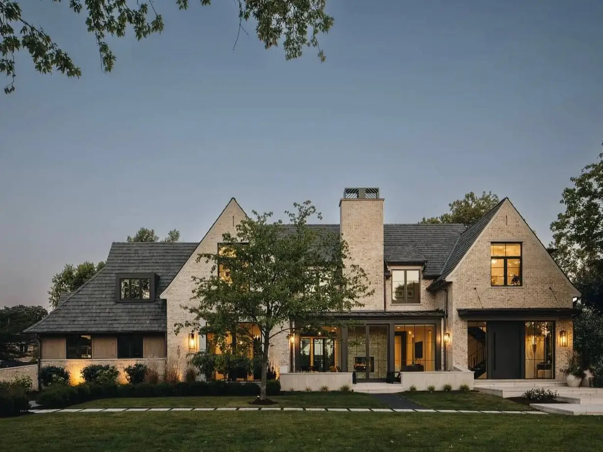 A large beige brick and black house exterior with a dark slate roof and warm interior lighting shown at dusk.