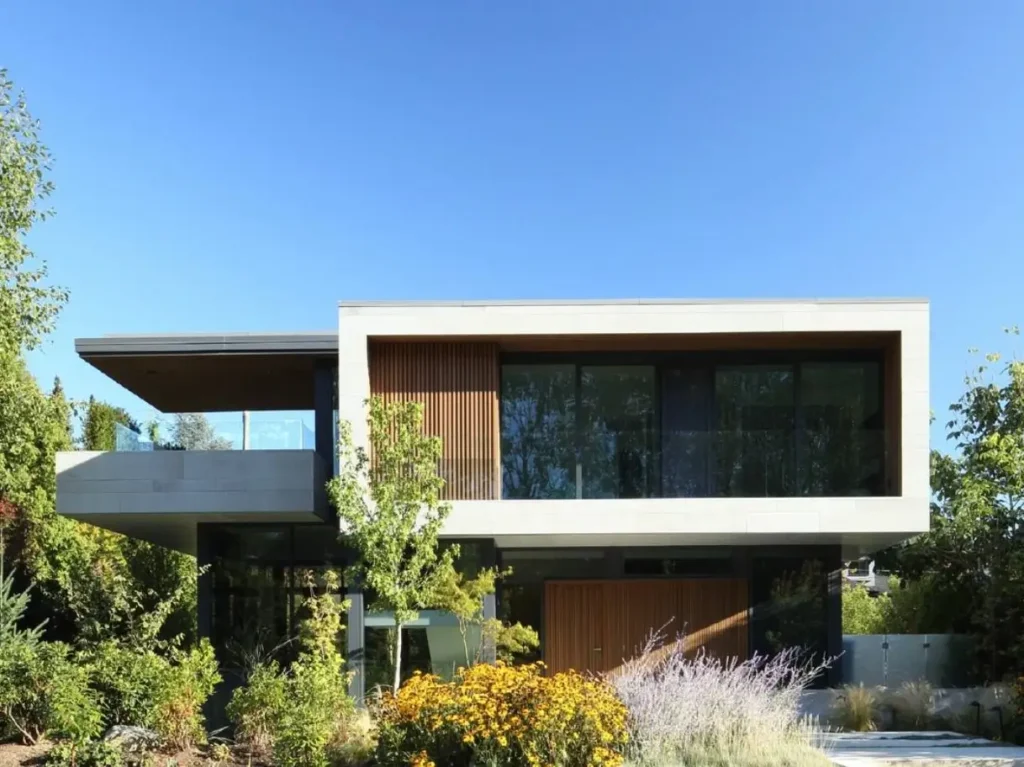 Exterior of a modern minimalist house design featuring a cantilevered upper level, wood accents, and large glass windows, set within a lush garden.
