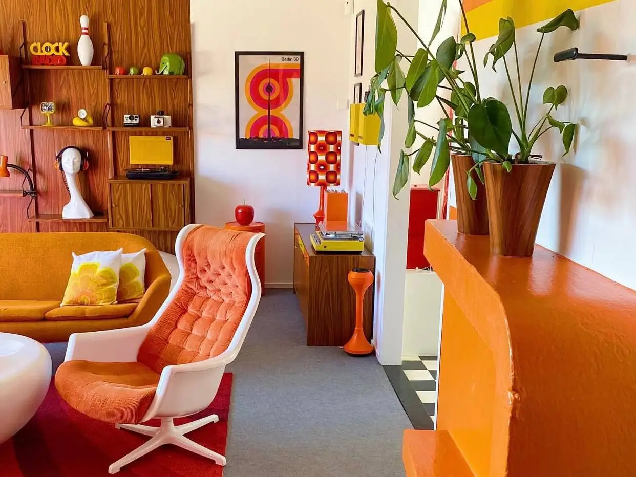 Living room showcasing retro 70s interior design style with orange lounge chair, yellow sofa, and teak wood shelving.