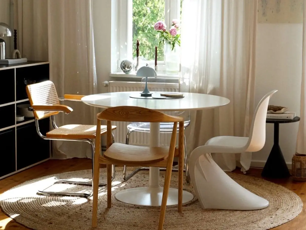 A sunlit condo dining nook featuring a round white pedestal table with a mix of modern and mid-century chairs on a jute rug.
