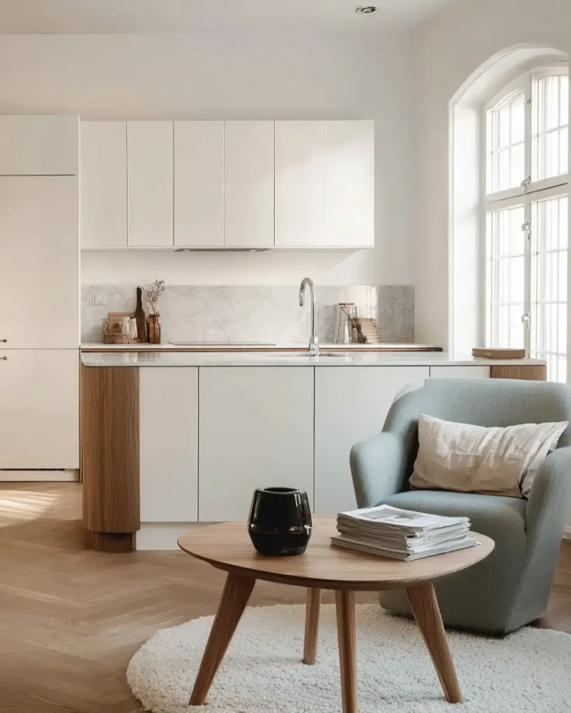 A contemporary Scandinavian kitchen nook with white cabinets, a marble backsplash, and a blue armchair on a herringbone wood floor.