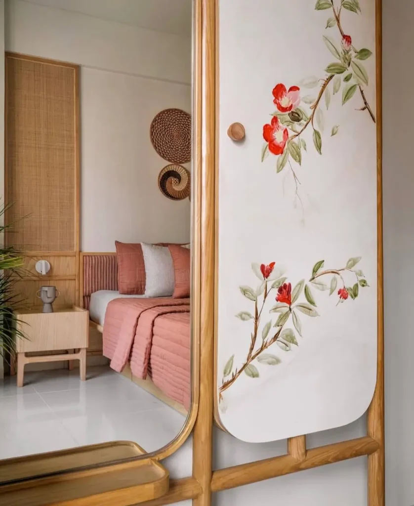 A reflection in a mirror shows a modern bedroom with a pink bed, rattan headboard, and a hand-painted floral design on a wooden door.