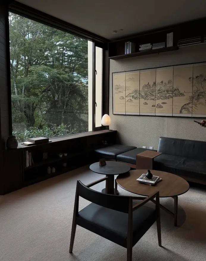 A moody Japandi living room with a large window framing a forest view dark furniture and a Japanese scroll.