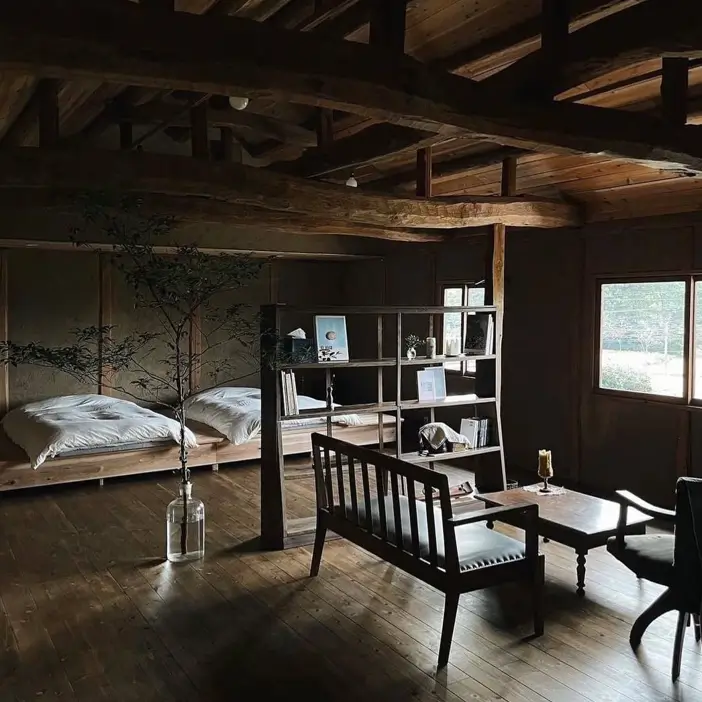 A rustic, moody Japanese-style room with dark exposed beams, two low beds, and a bookshelf room divider.