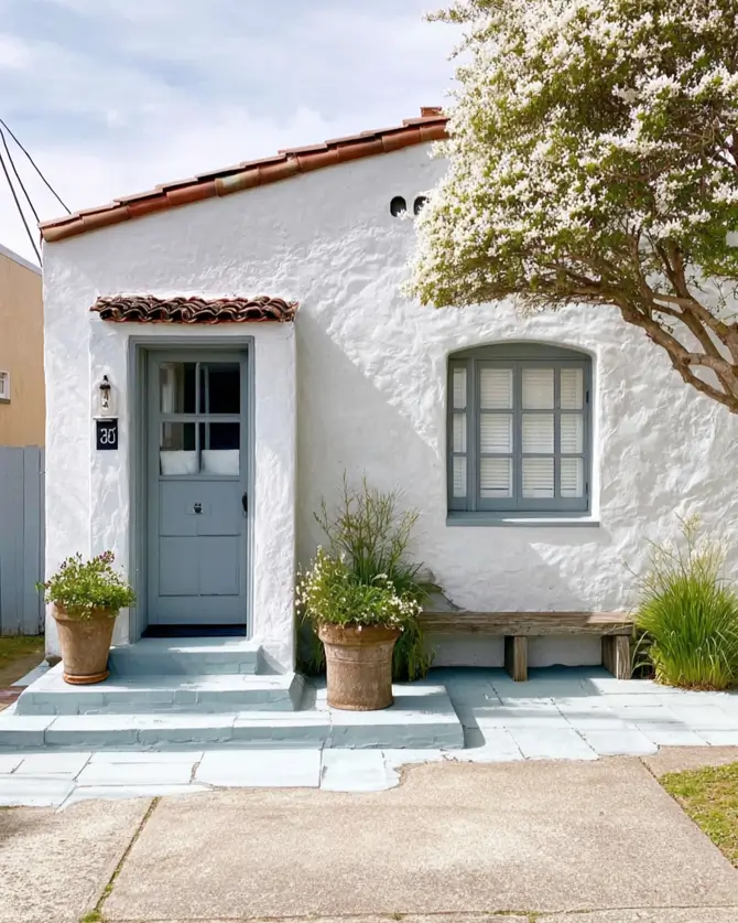 38 Small Mediterranean-Style Homes That Define Coastal Charm 20 White stucco small Mediterranean home cottage with soft grey-blue trim on the door, window, and front steps.